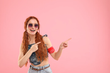 Stylish young hippie woman in sunglasses pointing at something on pink background, space for text