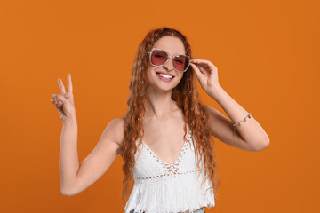 Stylish young hippie woman in sunglasses showing V-sign on orange background