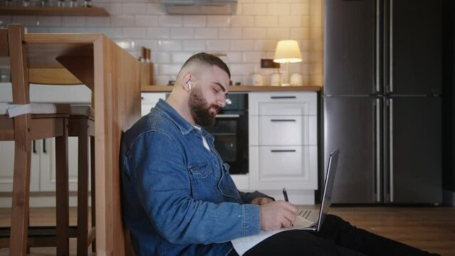 Young man student sitting on the floor leaning against the kitchen counter doing research working on laptop taking notes on paper while listening the music with in ear headphone
