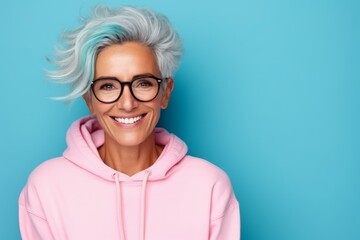 Portrait of smiling senior woman in pink hoodie and glasses on blue background - obrazy, fototapety, plakaty