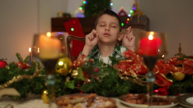 A boy, sitting at a festively decorated table for Christmas, makes wishes with his eyes closed and his fingers crossed