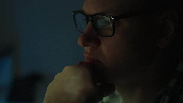 At night a man in glasses, who is an IT programmer, is busy working with a laptop computer. His serious face indicates deep analysis of the data on the screen. Reflection of information in his glasses