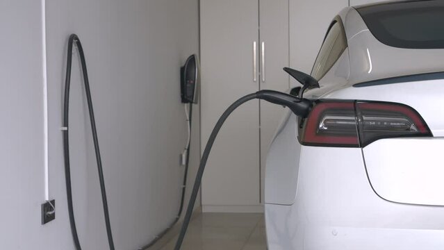 CLOSE UP: Black power cord connected to BEV for powering engine battery. White electric car plugged into a charging station while parked in home garage. Green alternative for modern transportation.