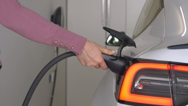 CLOSE UP: Woman plugs in a white BEV for charging motor battery in home garage. She connects black power cord after driving the electric vehicle. Effortless system for recharging an electric car.