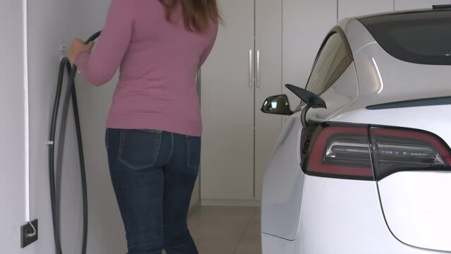 CLOSE UP: Lady unplugs and then sits into electric car to drive out of garage. Female driver unplugs black power cord from a BEV. An effortless system for an alternative and green means of transport.
