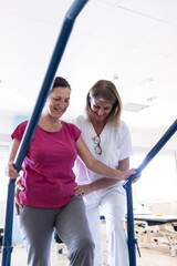 Vertical photo of a middle aged brunette woman in pink shirt. She is doing rehabilitation with her physiotherapist who is helping her.