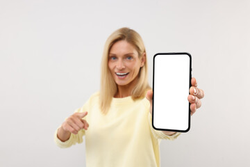 Happy woman holding smartphone and pointing at blank screen on white background, selective focus