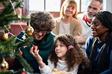 Multi-ethnic family and young daughter decorate Christmas tree in house. 