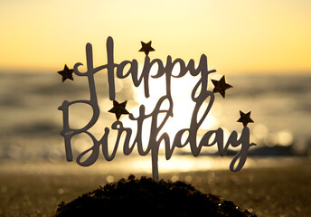 Stick in shape of letters Happy Birthday with stars at dawn and sunset on background of sea waves on sand seashore. Word Happy birthday in sandy beach on sunny summer day. Concept Happy Birthday