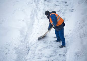 A man dressed in a uniform and an orange waistcoat clears a path from snow during a snowfall