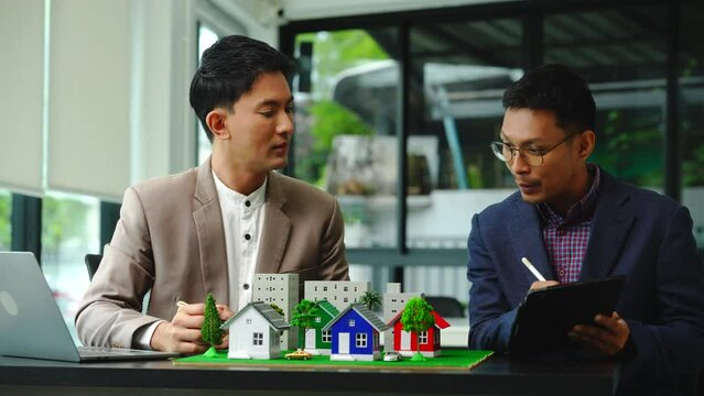 Two professionals smiling, discussing over model houses on a table, possibly planning or reviewing a housing project on large land. Asian people, only businessman, Middle-age