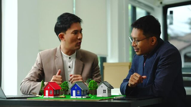 Two professionals smiling, discussing over model houses on a table, possibly planning or reviewing a housing project on large land. Asian people, only businessman, Middle-age