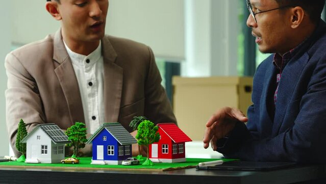 Two professionals smiling, discussing over model houses on a table, possibly planning or reviewing a housing project on large land. Asian people, only businessman, Middle-age