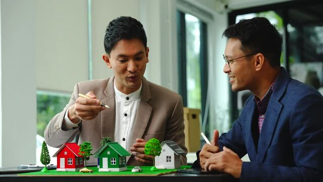 Two professionals smiling, discussing over model houses on a table, possibly planning or reviewing a housing project on large land. Asian people, only businessman, Middle-age