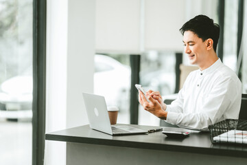 Fototapeta na wymiar Attractive smiling male asian business person in white shirt working on laptop at desk with coffee cup and documents nearby.
