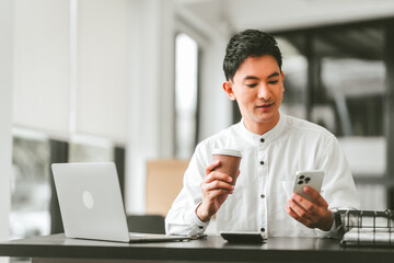 Attractive smiling male asian business person in white shirt working on laptop at desk with coffee cup and documents nearby.