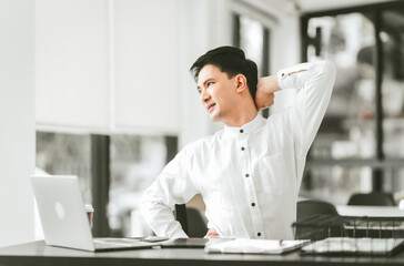 Asian middle age business man in white shirt stretching his neck, looks like he is having neck and back pain at his desk.