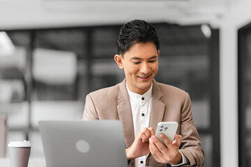 Asian professional male business person in beige suit and white shirt is working on laptop and holding pen, looking focused.