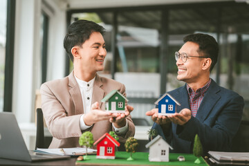 Two professionals smiling, discussing over model houses on a table, possibly planning or reviewing a housing project on large land. Asian people, only businessman, Middle-age