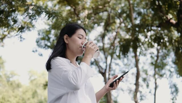 Young Asian woman using smartphone standing on a sunny day while drinking takeaway coffee in the park. Lifestyle and women concept.