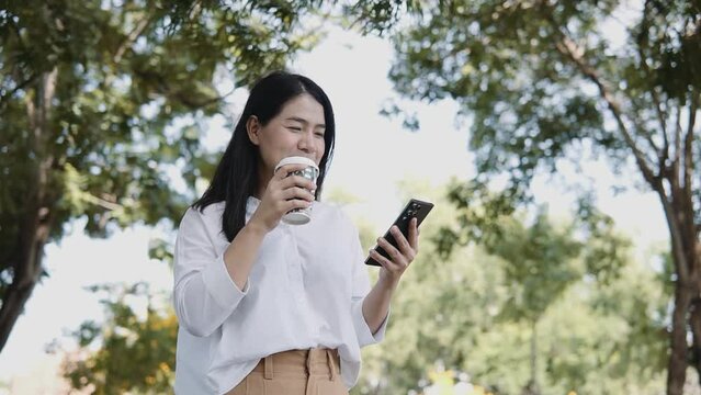 Young Asian woman using smartphone standing on a sunny day while drinking takeaway coffee in the park. Lifestyle and women concept.