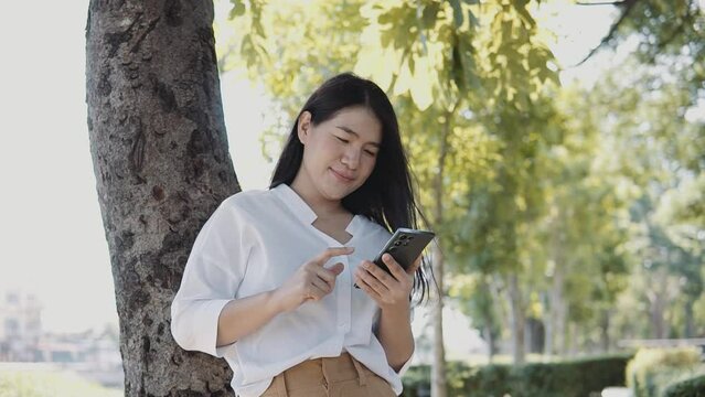 A Young Asian woman leaning on tree with a smartphone in the park .Enjoy and be happy. Lifestyle and women concept.