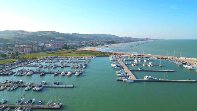 Inviting cinematic aerial push-in shot of the Port of Porto San Giorgio, with docked boats and the waters of the Adriatic Sea thriving in small sun-kissed ripples, larger waves crashing on the beach