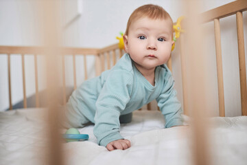 Fototapeta premium Close up of cute baby boy with blue eyes looking at camera while crawling in crib, copy space