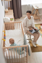 High angle portrait of smiling young mother working from home and using laptop with baby boy in...