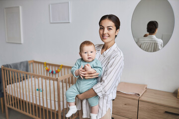 Waist up portrait of happy young mother holding baby son in cozy room, both looking at camera