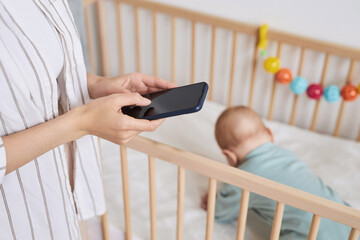 Close up of unrecognizable mother holding smartphone with blank screen while caring for baby boy...
