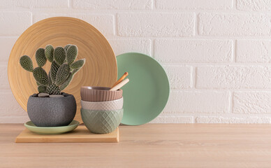 Ceramic bowls, dishes and tray on a wooden kitchen countertop, a cactus in a stylish pot in the...