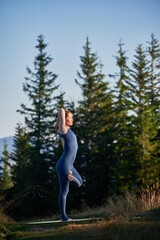 Middle-aged woman doing eagle pose. Young female practicing yoga poses in coniferous forest. Woman in dark blue tracksuit exercising in morning.