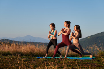 Pretty girls doing yoga at sunrise. Young mother meditating with her daughters in mountains. Outdoor workout concept. - obrazy, fototapety, plakaty