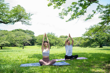 A beautiful Asian couple pull out yoga mats and practise yoga in a lush green grass park on a sunny day. Healthy living, mind-body care, urban living, autonomy, happy lifestyle - Powered by Adobe