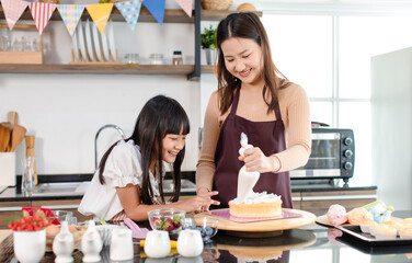 Asian beautiful female baker pastry chef mother wears apron standing smiling helping teaching little girl daughter decorating cake with whipping cream making homemade bakery in decorated home kitchen