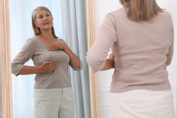 Beautiful senior woman doing breast self-examination near mirror indoors