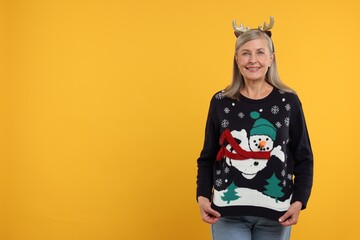 Happy senior woman in deer headband showing Christmas sweater on orange background. Space for text