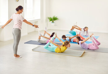 Happy children girls doing stretching exercises together lying on the floor on yoga mat in studio with female trainer. Group of kids doing dance sport workout in choreography class.