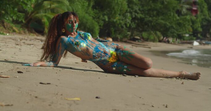 Sexy girl in body paint laying on the sand with ocean waves in the background on a tropical island