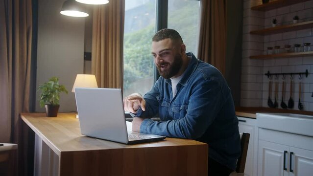 Smiling young adult man sitting against the kitchen counter having video call conversation through using laptop. Share news, talk to family, enjoy webchat virtual meeting