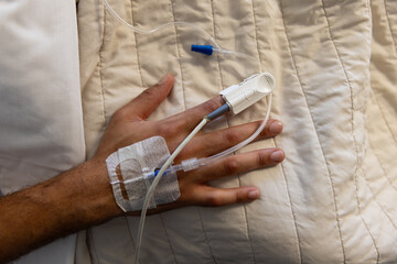 Hand of biracial male patient with iv drip lying in bed in sunny hospital room