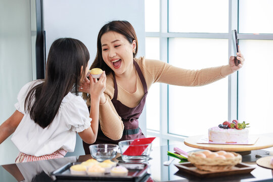 Asian beautiful female baker pastry chef mother using smartphone taking selfie photo with little girl daughter wearing apron standing smiling holding cupcakes posing together after finish baking cake