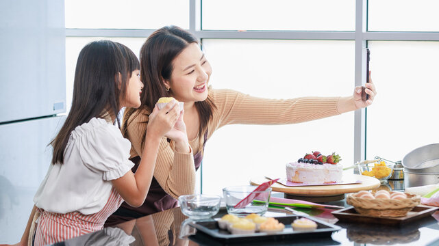 Asian beautiful female baker pastry chef mother using smartphone taking selfie photo with little girl daughter wearing apron standing smiling holding cupcakes posing together after finish baking cake