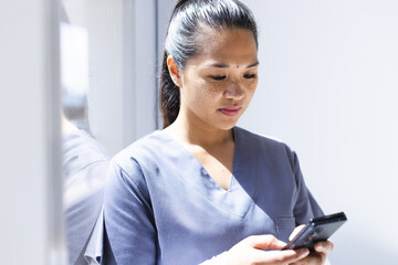 Biracial female doctor wearing scrubs using smartphon in hospital