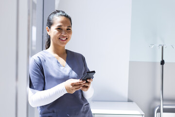 Portrait of biracial female doctor wearing scrubs using smartphon in hospital, copy space