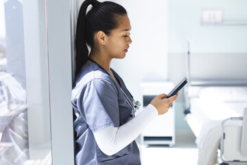 Biracial female doctor wearing scrubs using smartphon in hospital