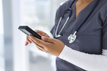 Midsection of biracial female doctor wearing scrubs using smartphon in hospital