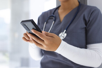 Midsection of biracial female doctor wearing scrubs using smartphon in hospital