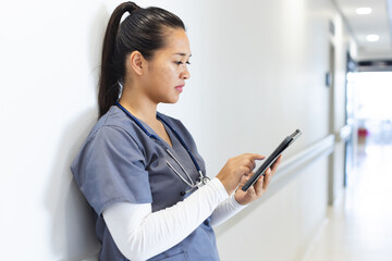 Biracial female doctor wearing scrubs using tablet in hospital corridor